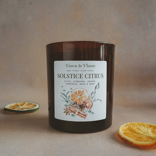 Fawn & Flame Solstice Citrus hand poured vegan candle in amber glass jar against hazy lavender backdrop and dried oranges. Label shows watercolour illustration of oranges, cinnamon, cloves and eucalyptus.