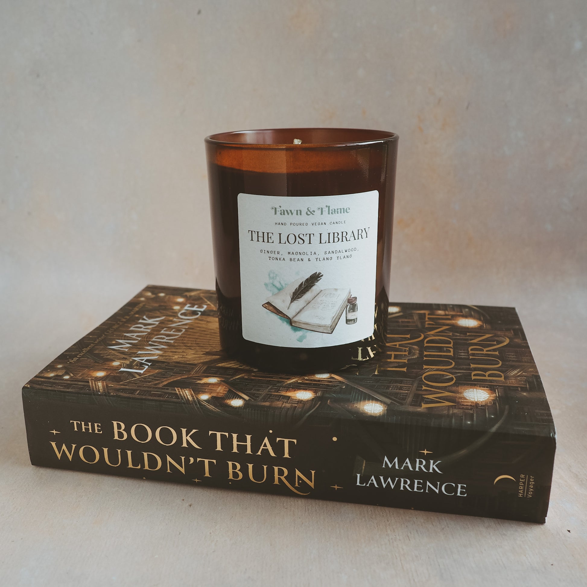 Fawn & Flame The Lost Library hand poured vegan candle in amber glass jar against hazy lavender backdrop with a book underneath, The Book That Wouldn't Burn by Mark Lawrence. Label shows watercolour illustration of an old book, quill and ink.