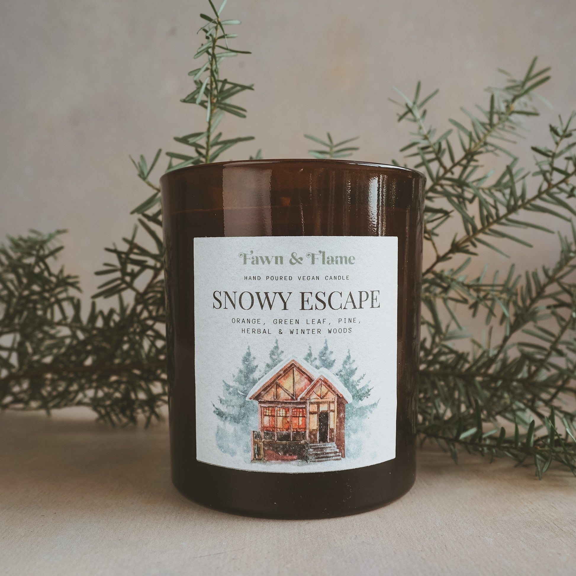 Fawn & Flame Snowy Escape hand poured vegan candle in amber glass jar against hazy lavender backdrop and pine. Label shows watercolour illustration of oranges, cinnamon, cloves and eucalyptus.