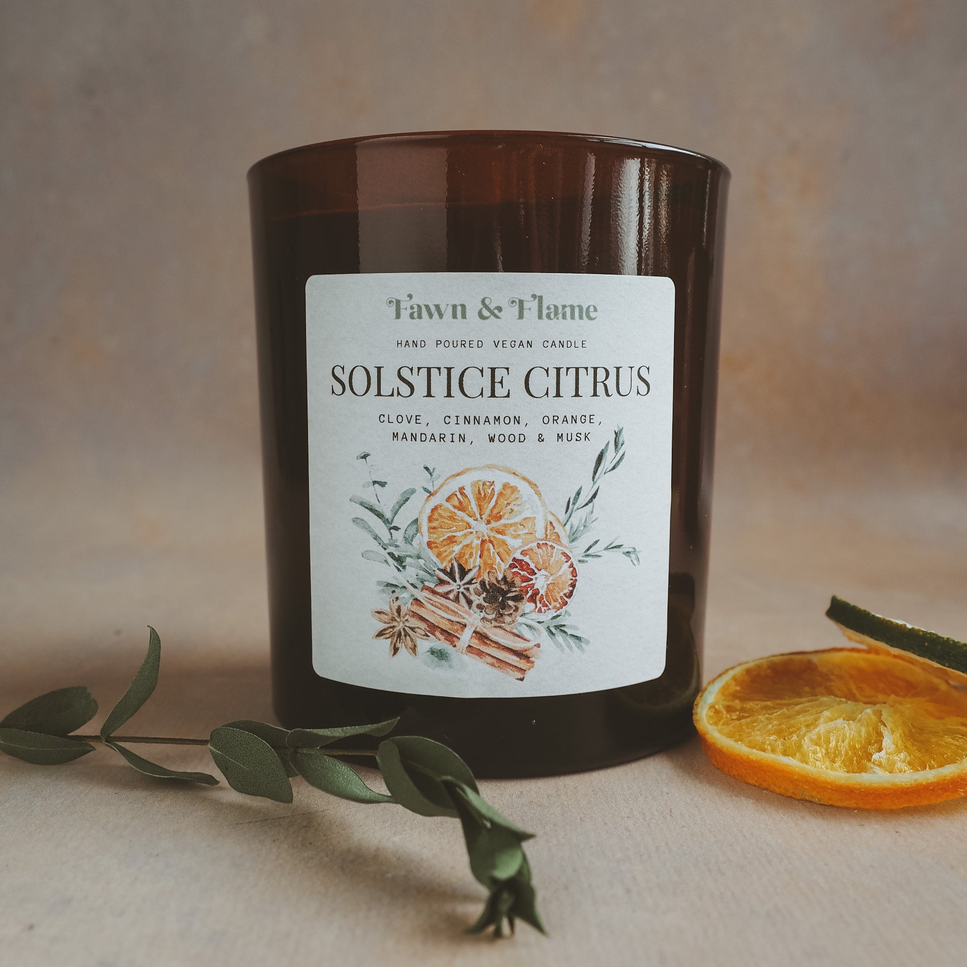 Fawn & Flame Solstice Citrus hand poured vegan candle in amber glass jar against hazy lavender backdrop and dried oranges and greenery. Label shows watercolour illustration of oranges, cinnamon, cloves and eucalyptus.