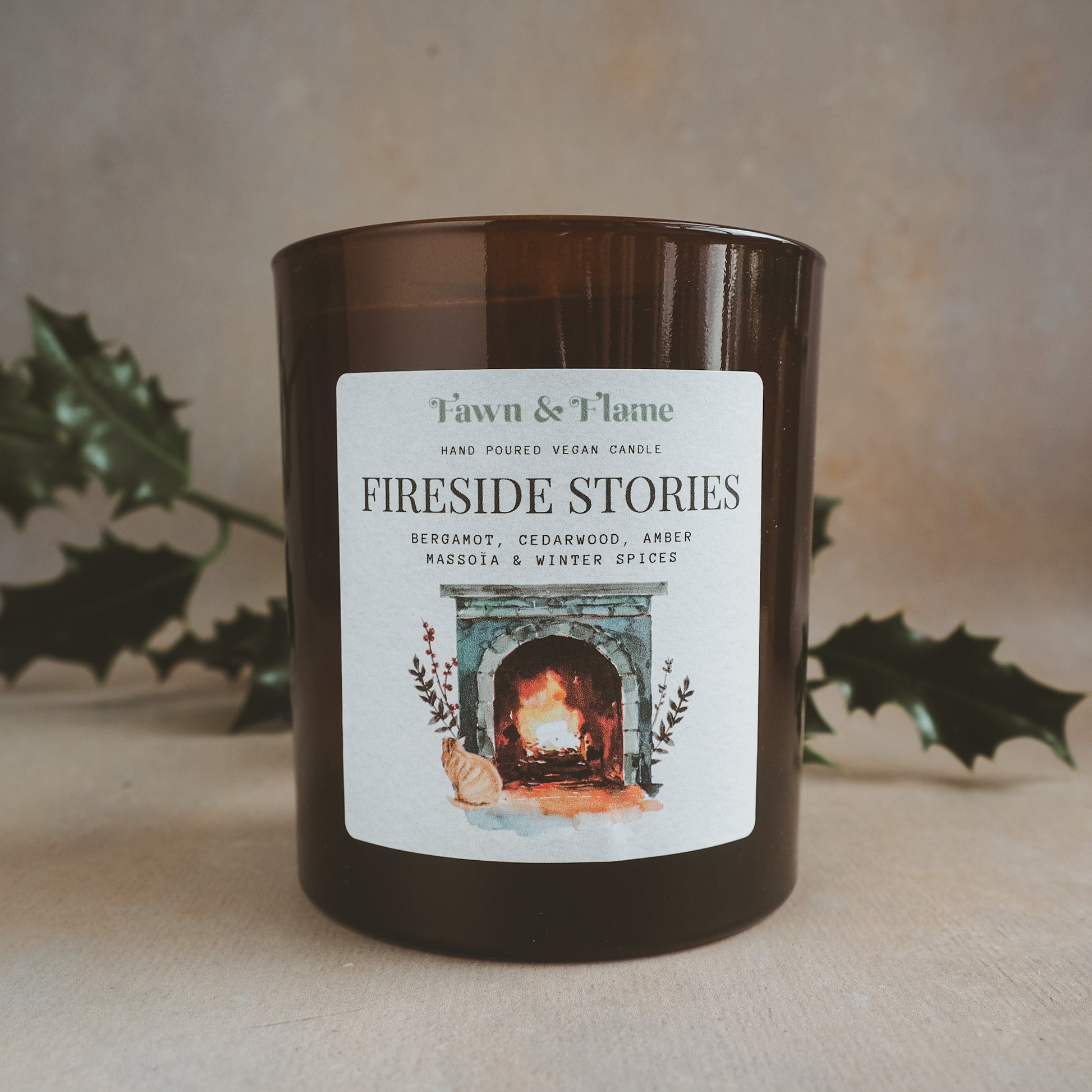 Fawn & Flame fireside stories hand poured vegan candle in amber glass jar against hazy lavender backdrop and sprig of holly. Label shows watercolour image of an old stone fireplace, with a ginger cat in front of it.