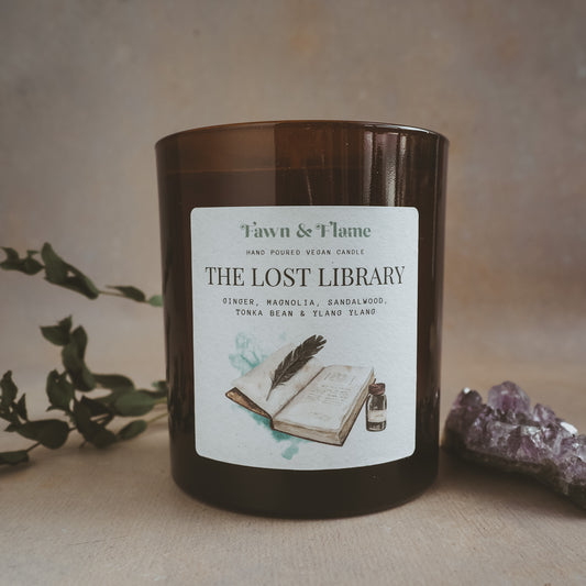 Fawn & Flame The Lost Library hand poured vegan candle in amber glass jar against hazy lavender backdrop with dried eucalyptus and amethyst crystal. Label shows watercolour illustration of an old book and ink.